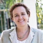 Dr Briony Scott smiles at the camera, wearing a grey linen jacket, earrings, and a silver necklace.